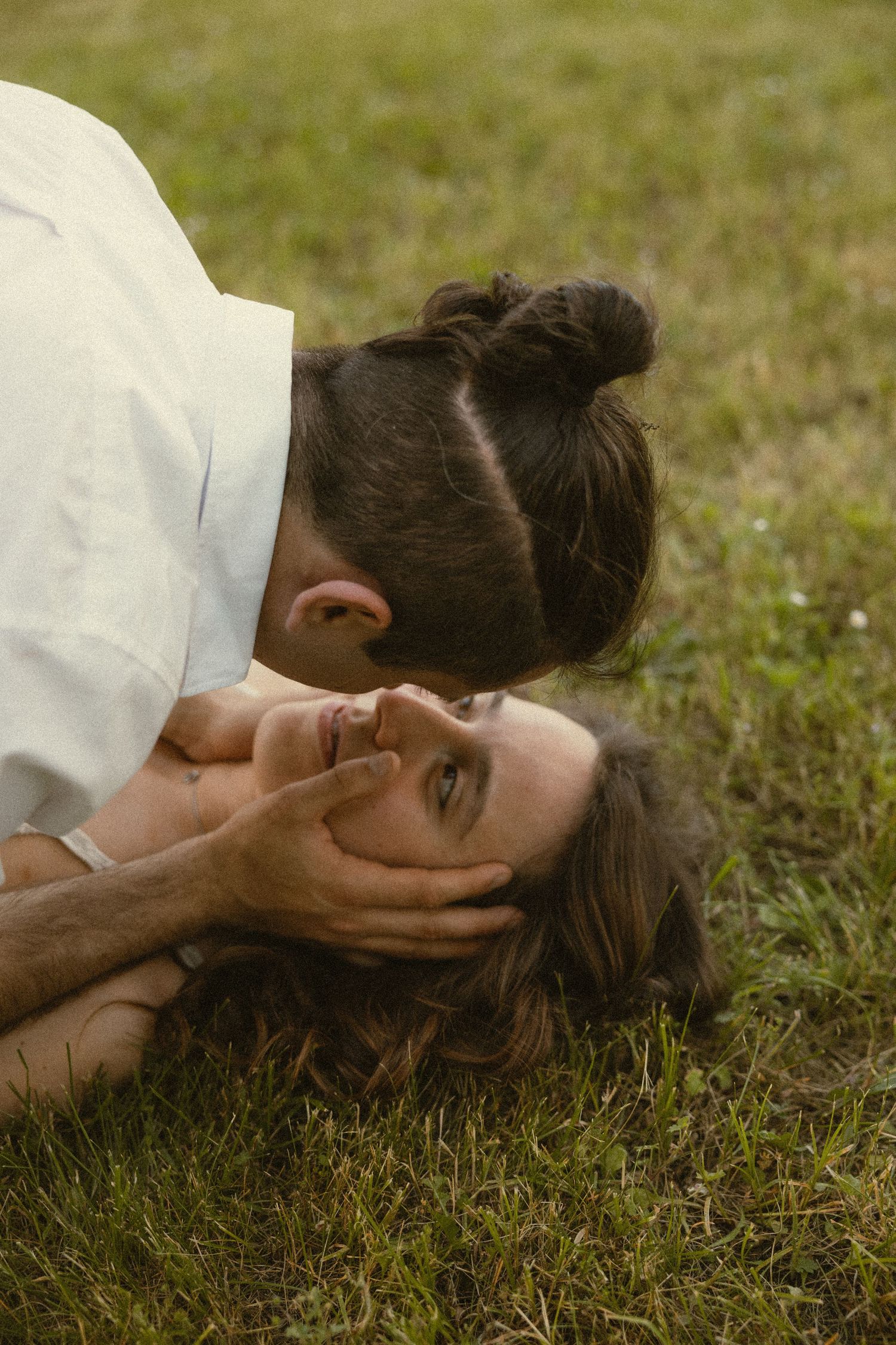 Couple lying on green grass as a man with a bun leans over to kiss a smiling woman while cradling her face in an intimate outdoor moment.