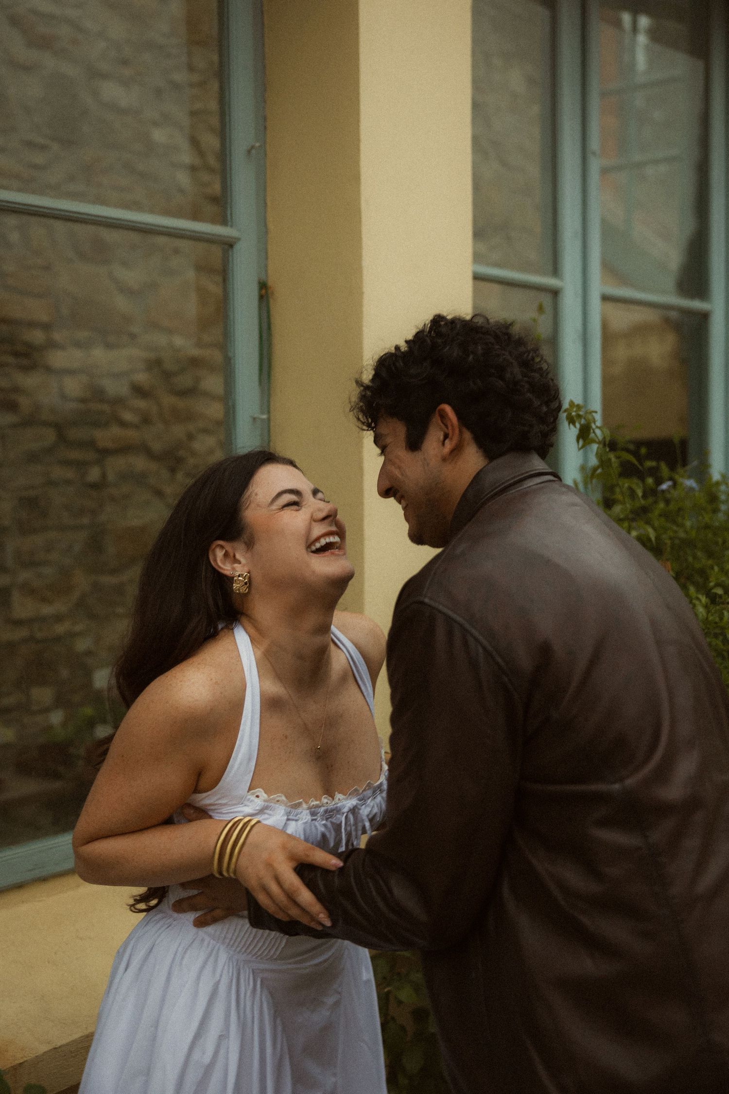 A laughing woman in a white sundress holds hands with a curly-haired man in a leather jacket outside a stone building.