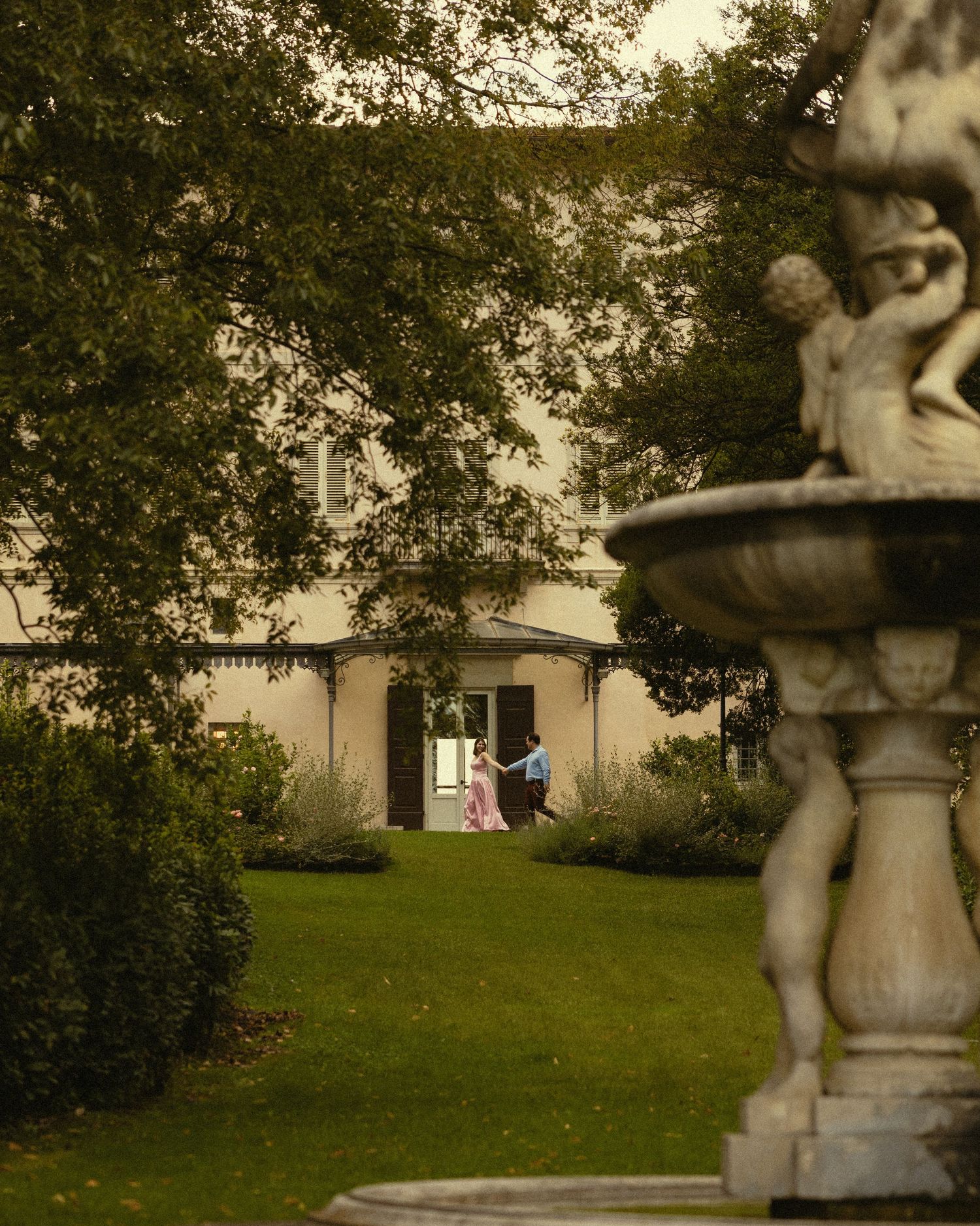 Stone fountain with cherub statues in the foreground overlooking a green lawn and a cream-colored historic house framed by tall trees.