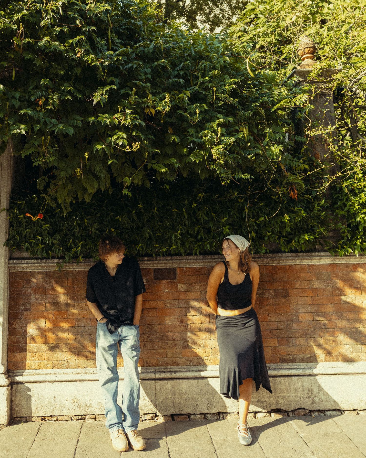 A man and a woman lean against a sunlit brick wall topped with lush green ivy while standing on a stone sidewalk in Italy.
