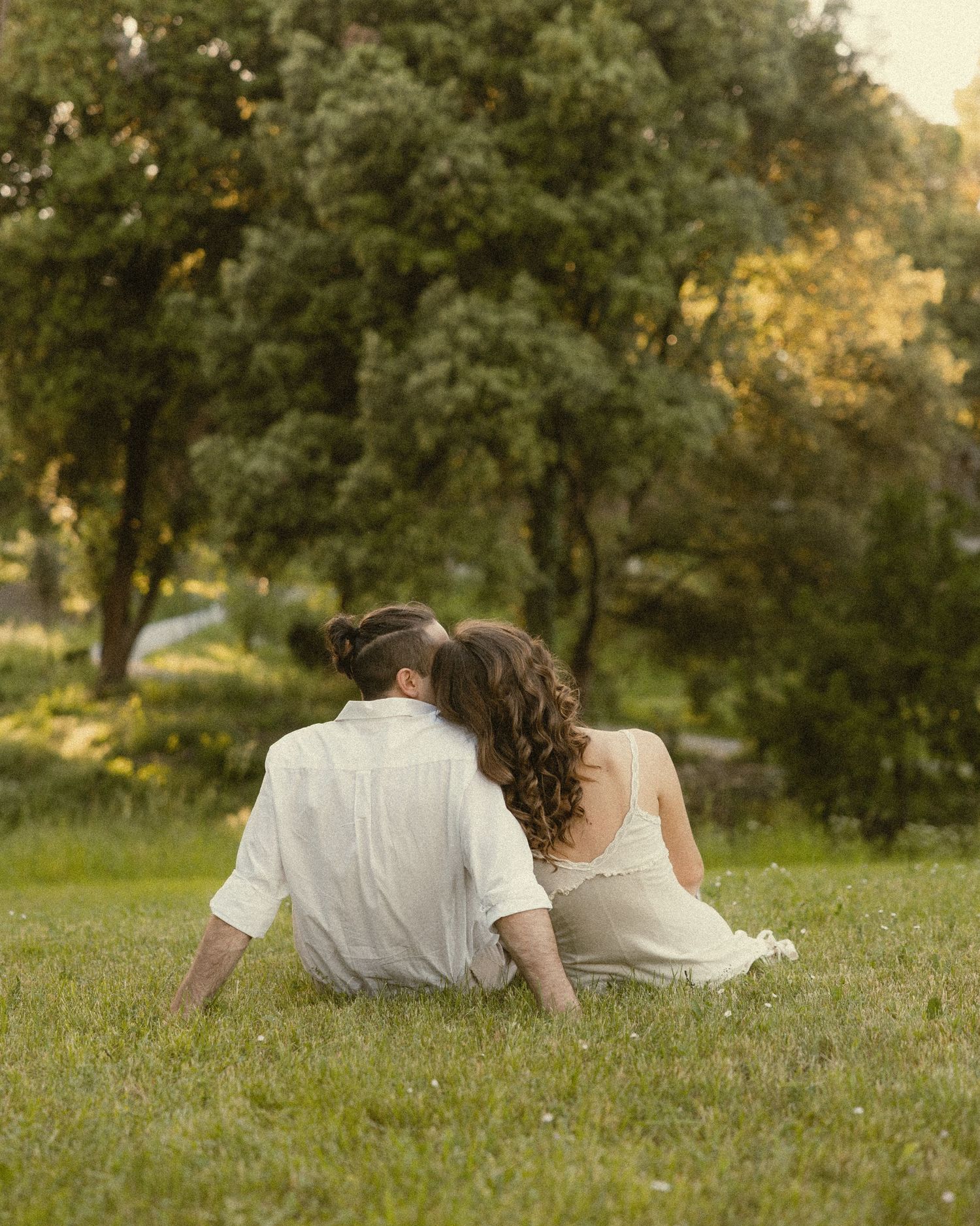 Couple sitting close together on a sunny grassy hill, seen from behind, with lush green trees in the background.