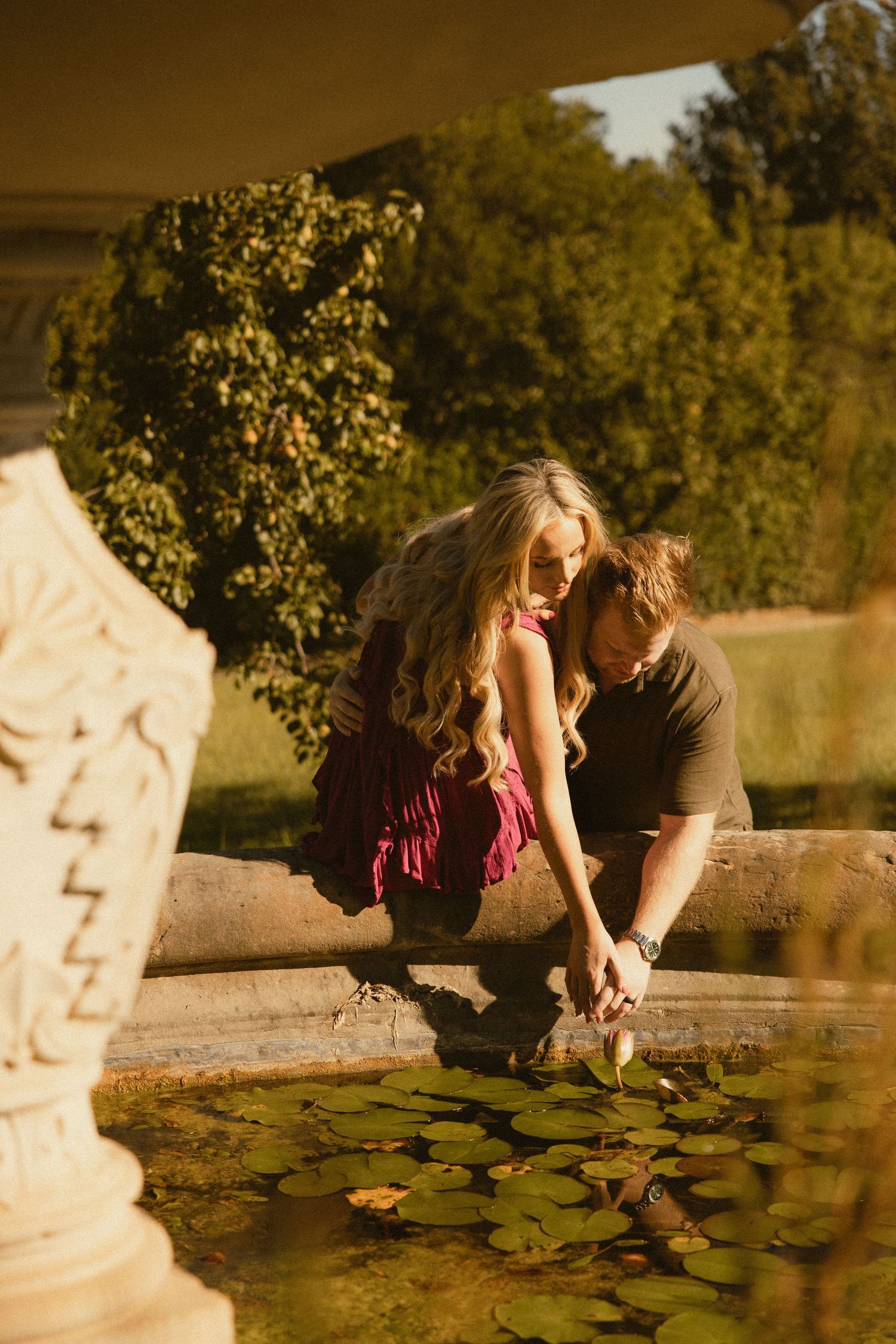Couple sitting on a stone ledge reaching toward lily pads in a garden pond, framed by lush green trees in warm golden sunlight.