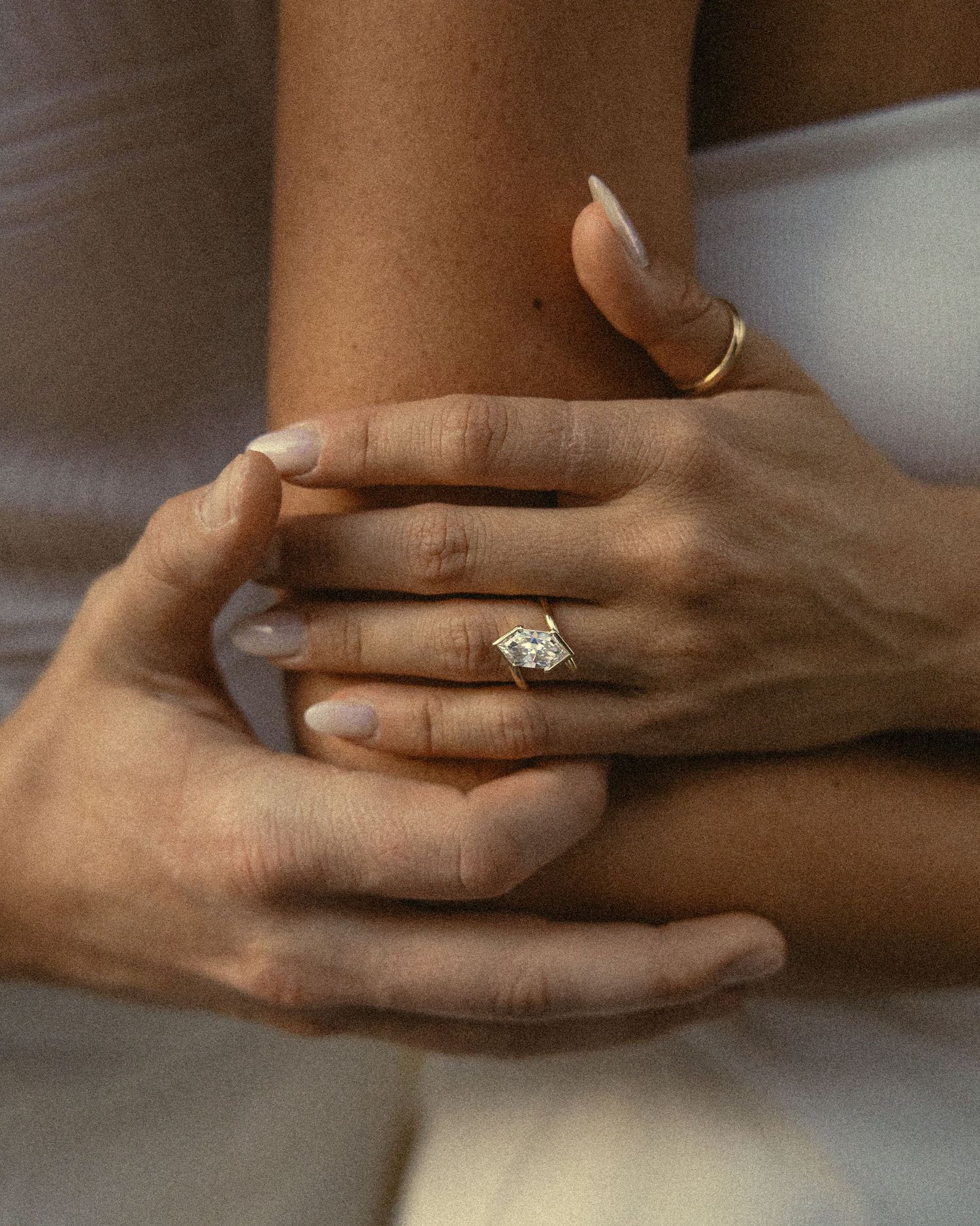 Close-up of two embracing hands, one wearing a marquise-cut diamond engagement ring and the other a thin gold band, with soft neutral tones.
