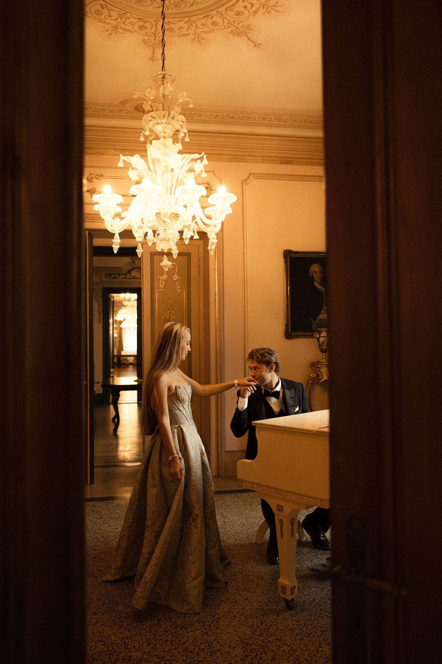 Elegant woman in a flowing gown extends her hand to a seated man in a tuxedo at a white piano beneath a glowing crystal chandelier.