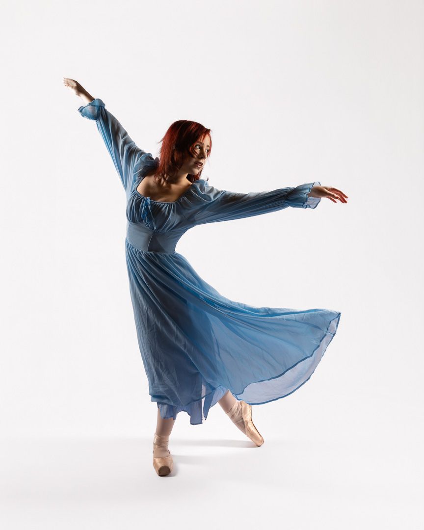 A graceful ballerina in a flowing blue dress performs an elegant pose on pointe shoes against a white background.