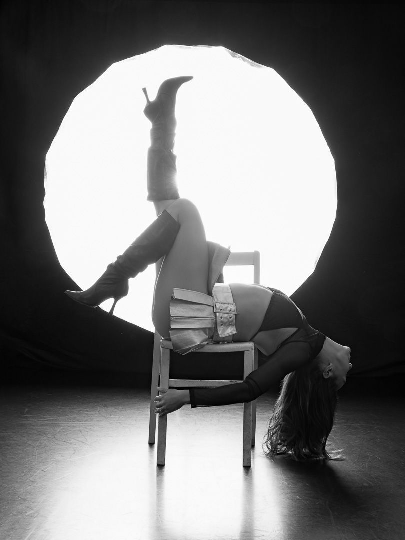 Black and white photo of a performer doing an acrobatic pose on a chair in front of a circular light backdrop.