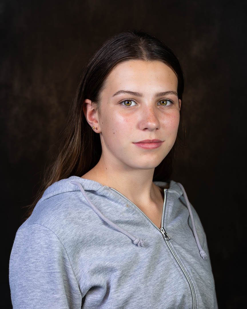 Young woman with brown hair wearing a gray zip-up hoodie poses for a portrait against a dark background.