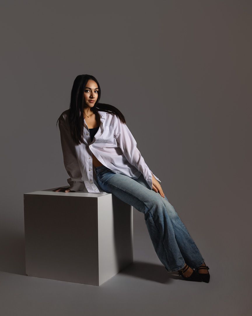 A model in a white button-down shirt and jeans poses on a white cube against a gray studio backdrop.