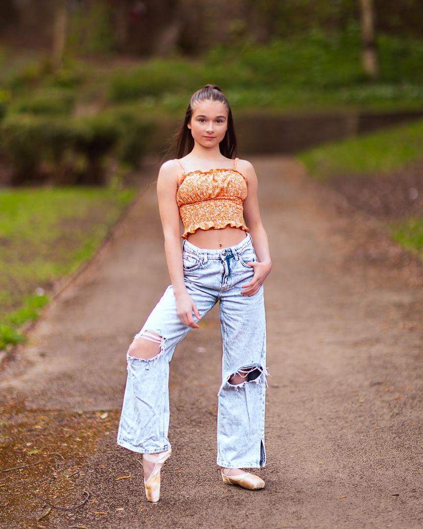 A young dancer poses in ripped jeans and orange crop top on a path, wearing ballet pointe shoes outdoors.