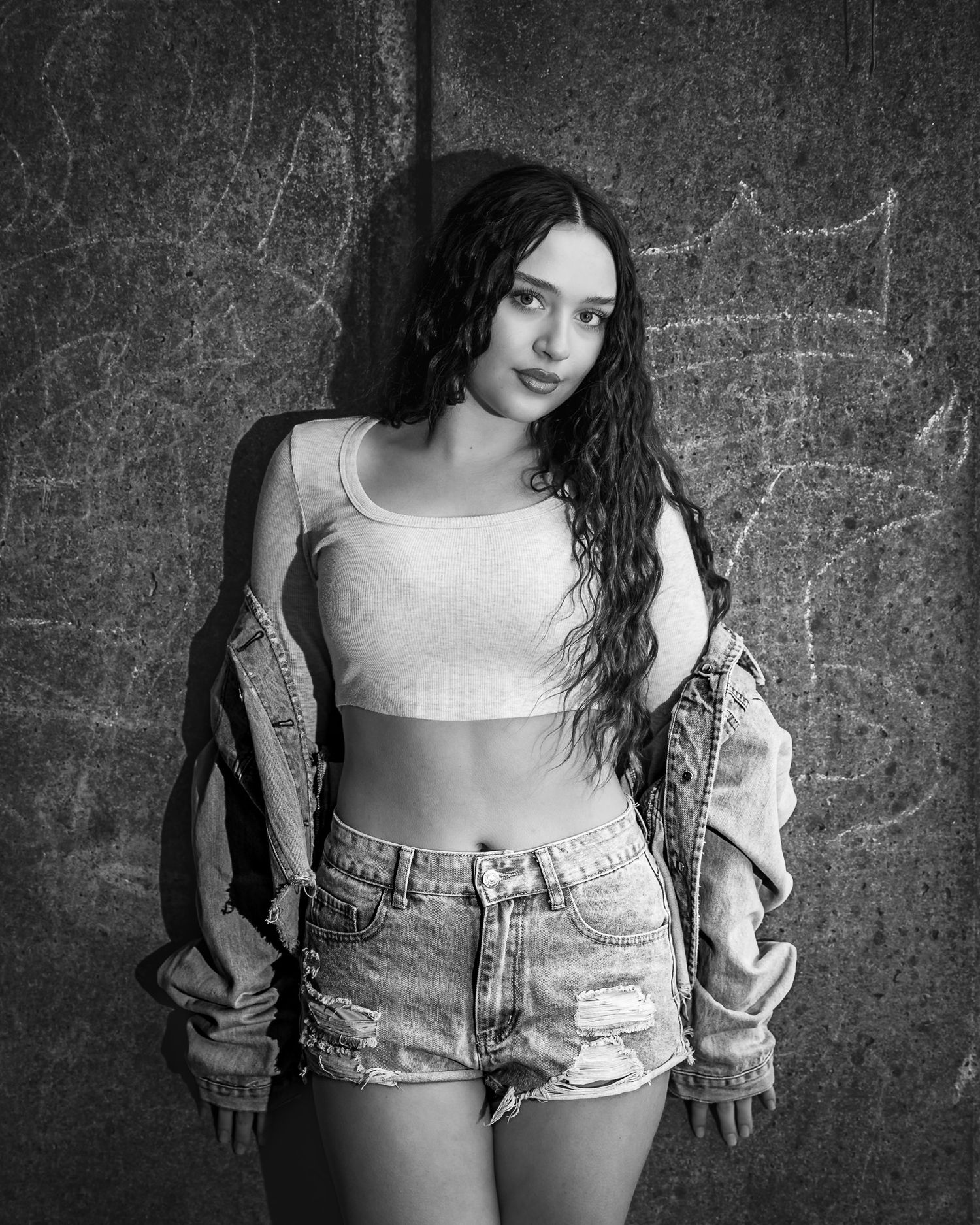 Black and white photo of a young woman in denim jacket and shorts leaning against a textured wall.