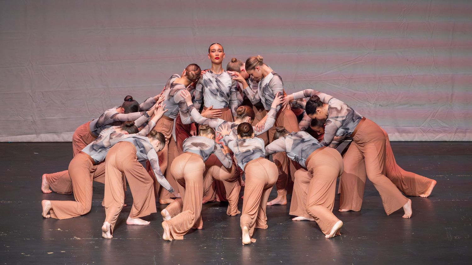 Dancers in a dramatic group pose on stage, with one performer standing center while others kneel around her in pink costumes.