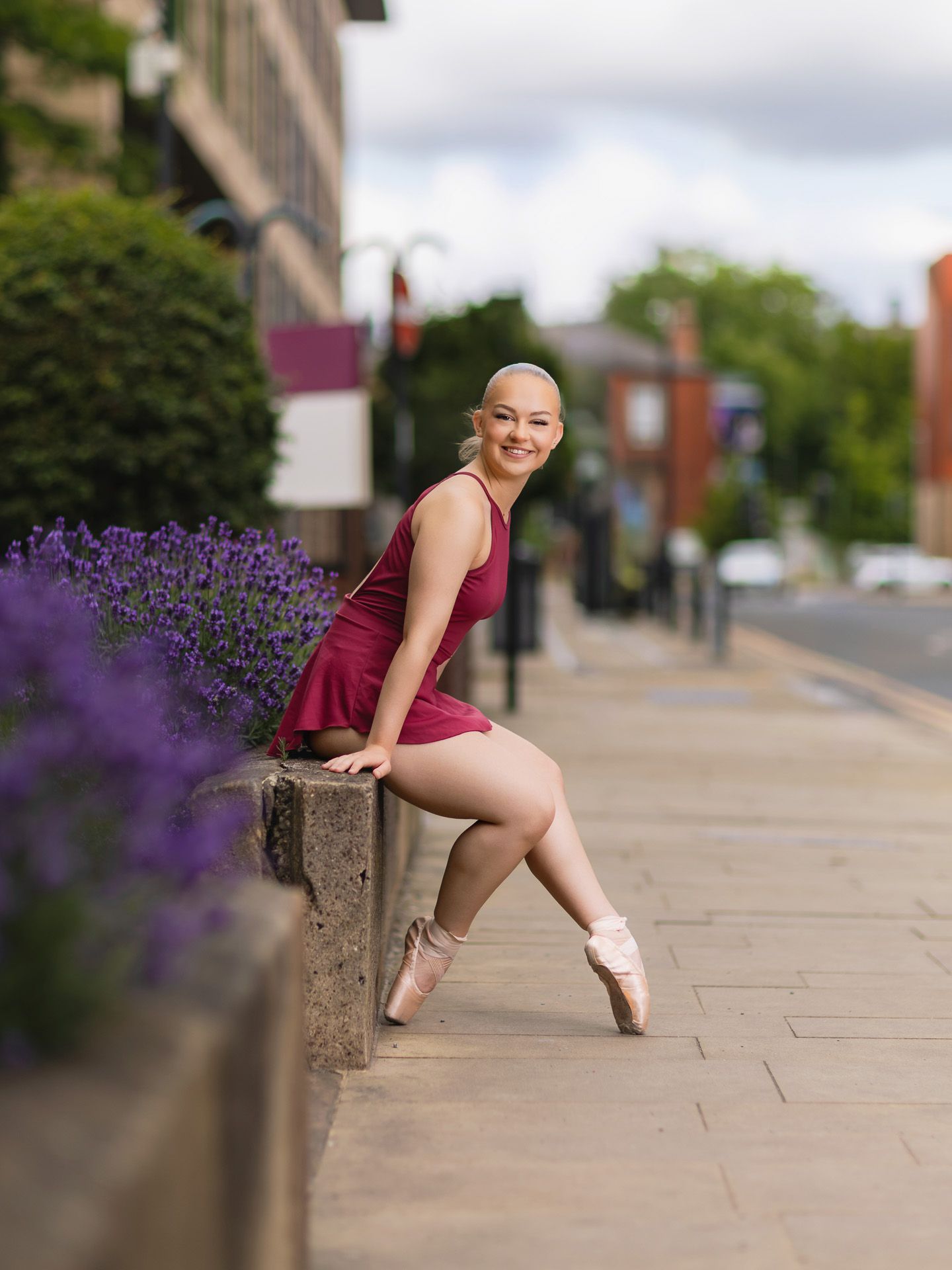 Smiling ballerina in burgundy dress poses en pointe on a sunny sidewalk with purple flowers.