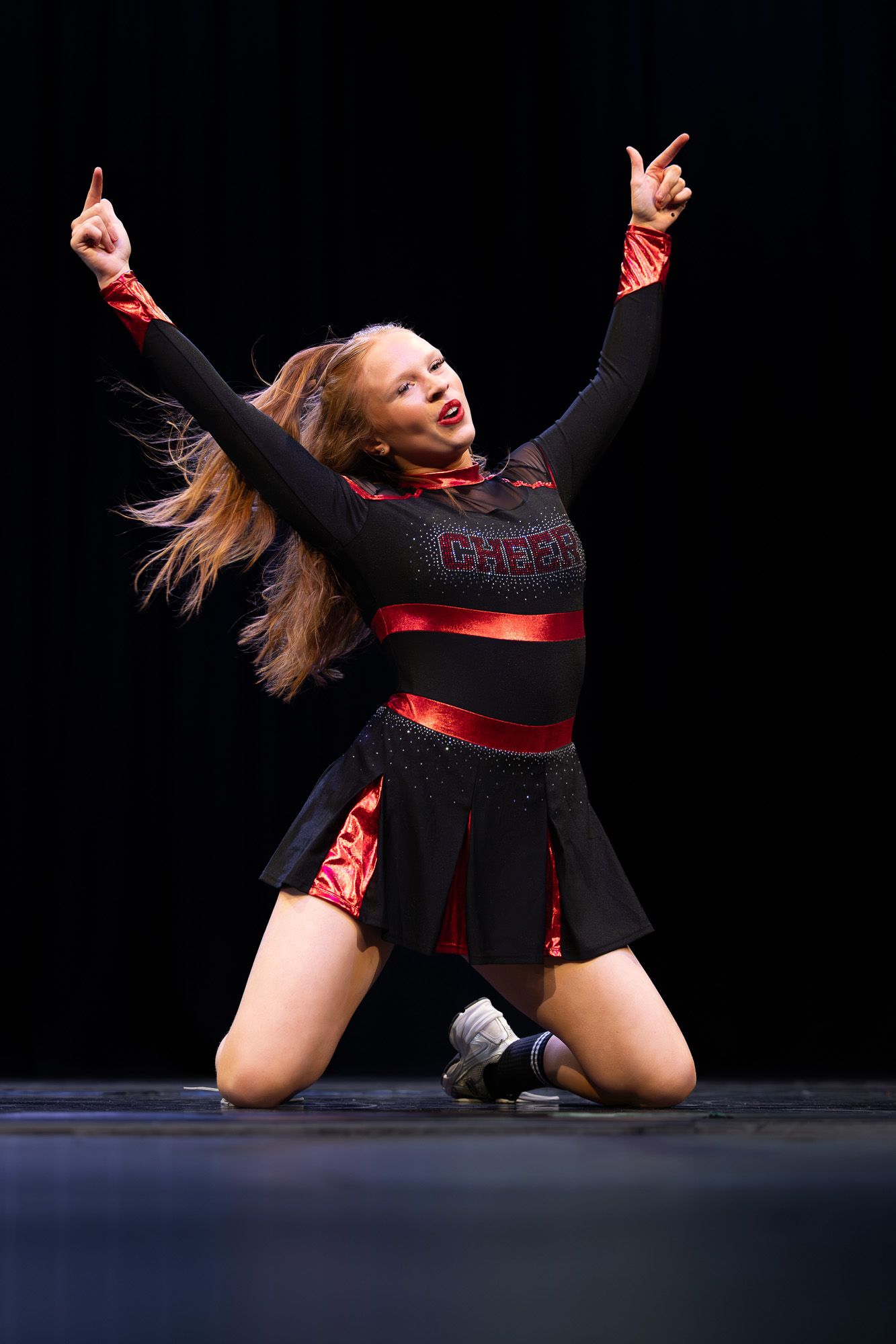A cheerleader in a black and red uniform kneels on stage with arms raised, performing an energetic routine under stage lights.