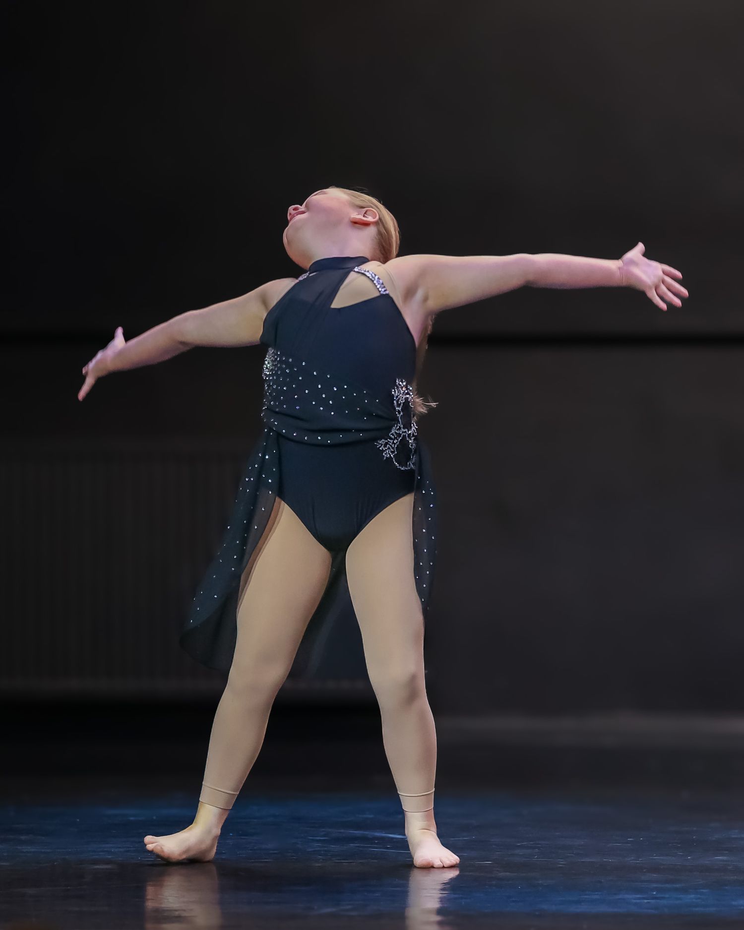 Young dancer in black leotard performs graceful pose with arms outstretched on stage.