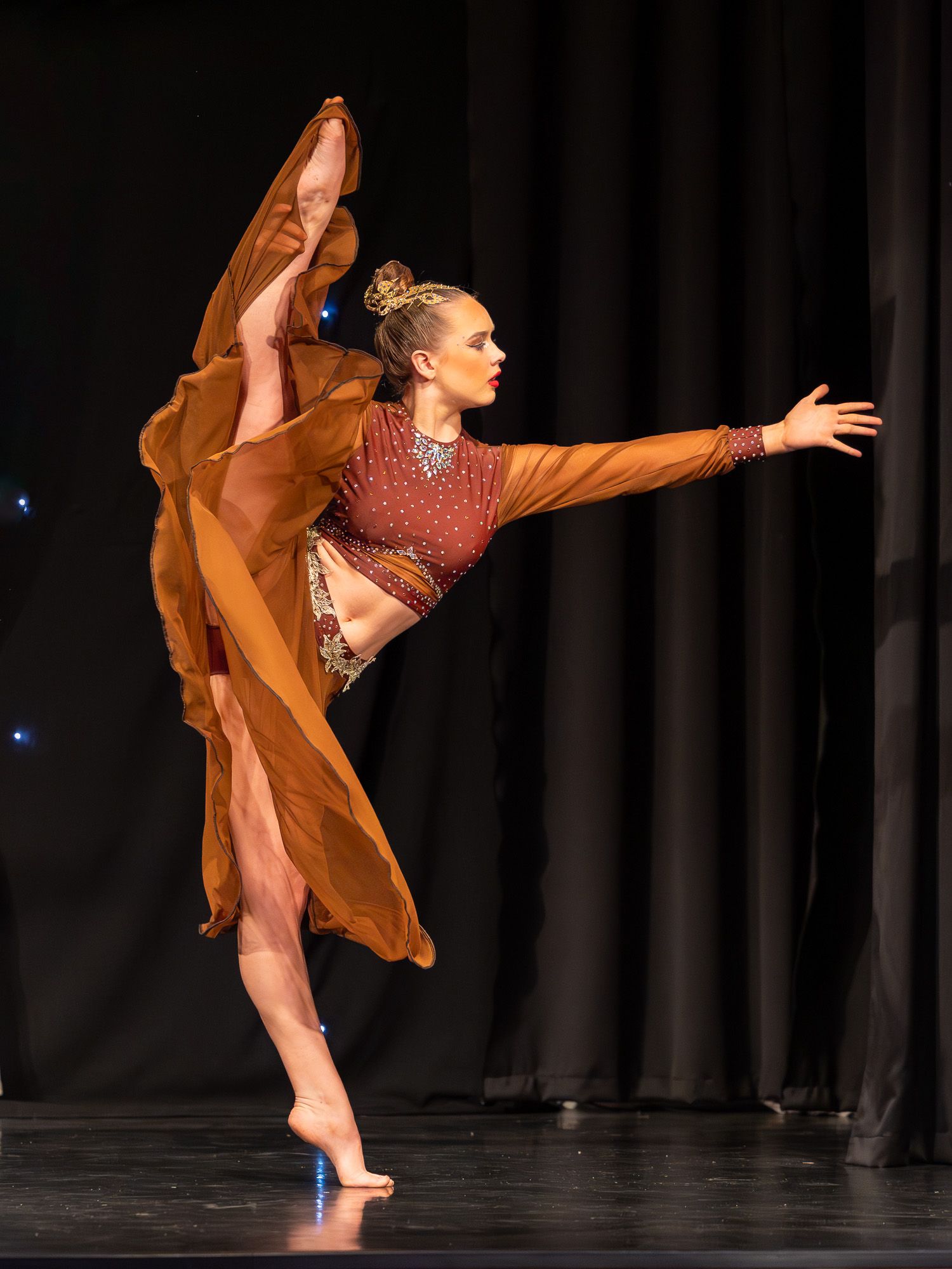 Graceful dancer in rust orange flowing dress performs an arabesque on stage under dramatic lighting.