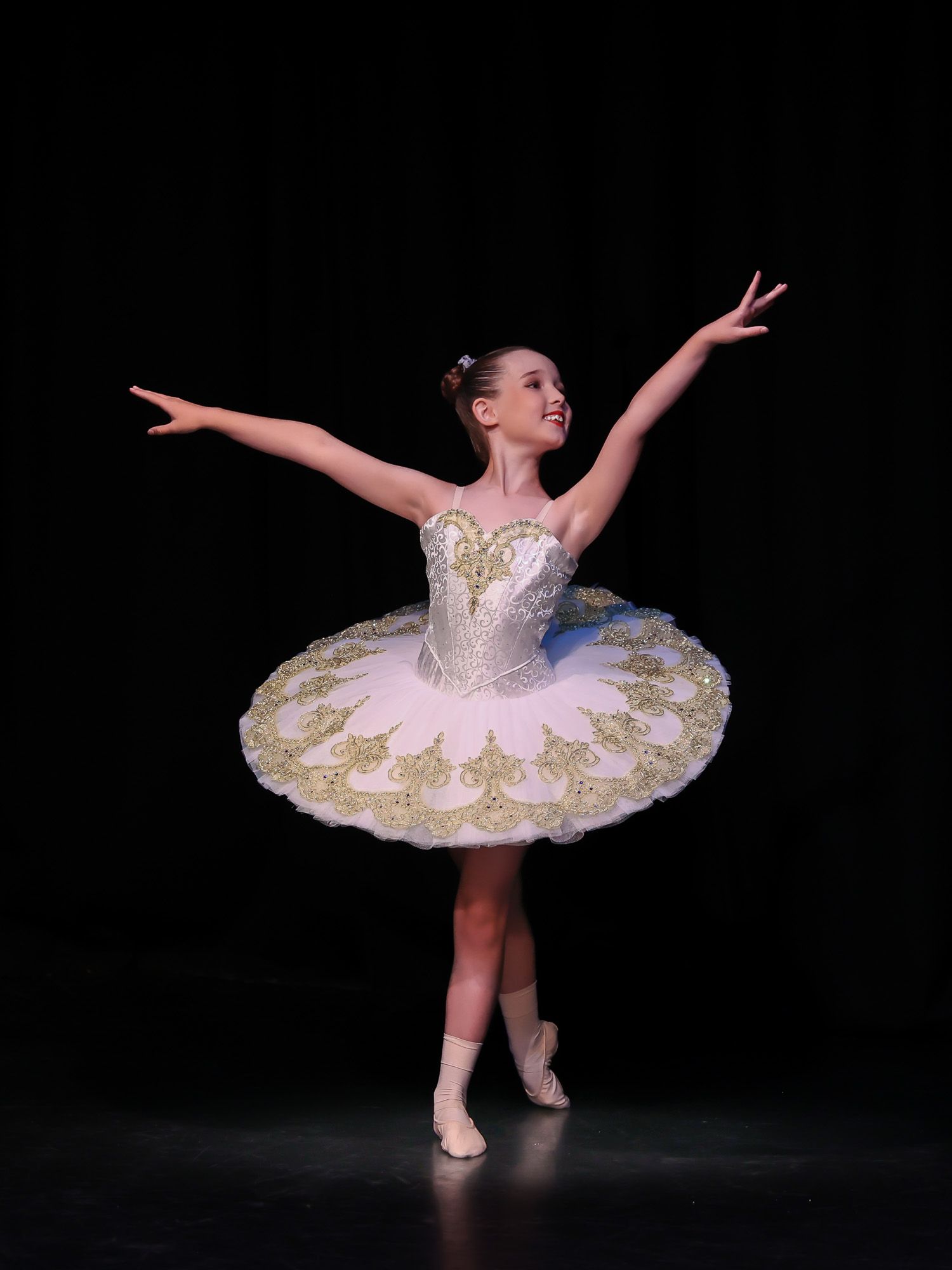 Young ballerina in white and gold tutu performs graceful arabesque on stage under spotlight.