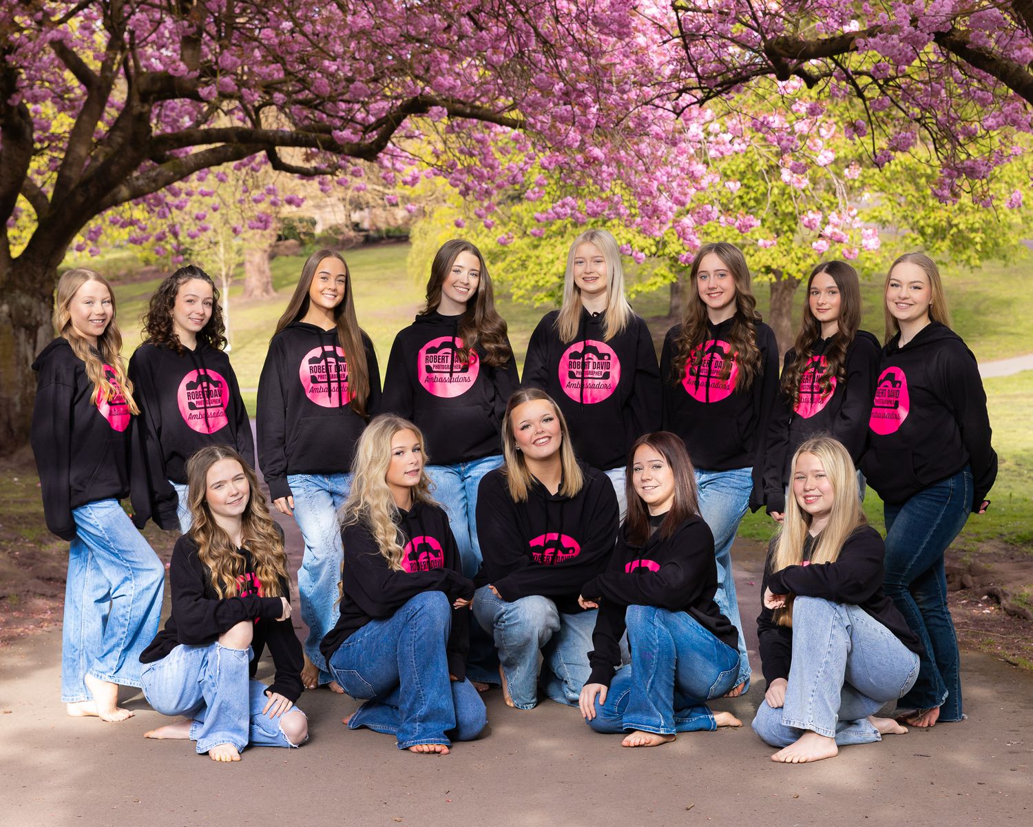 Group of teenage girls in matching black and pink hoodies posing under blooming pink cherry blossom trees outdoors.