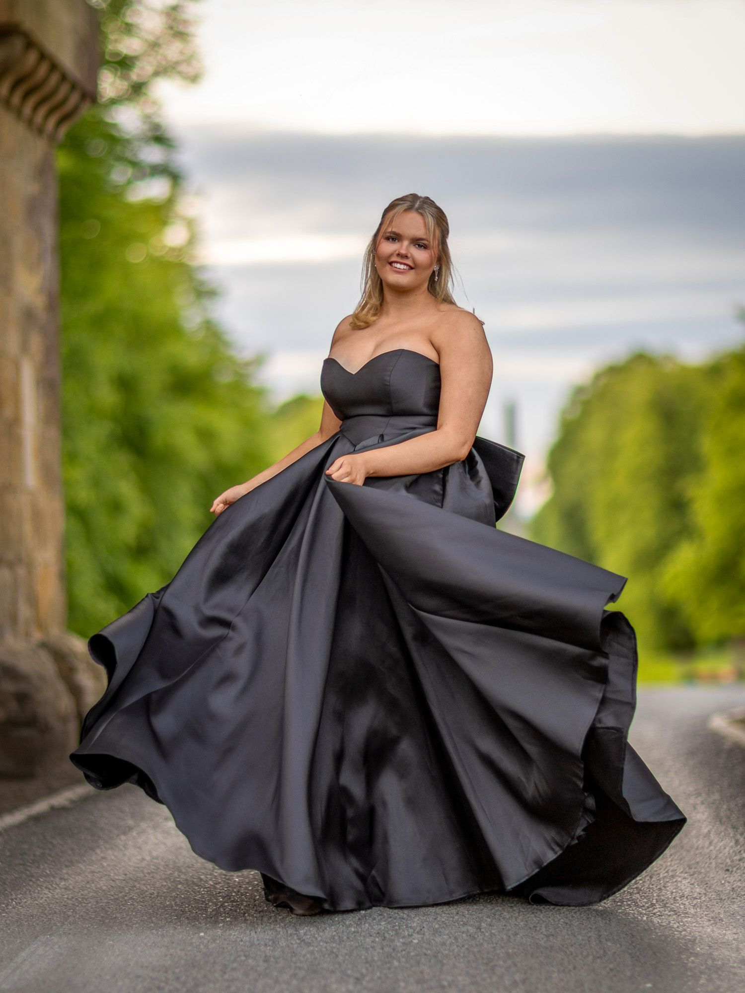 A dramatic black ballgown swirls on a tree-lined path during an outdoor photoshoot.