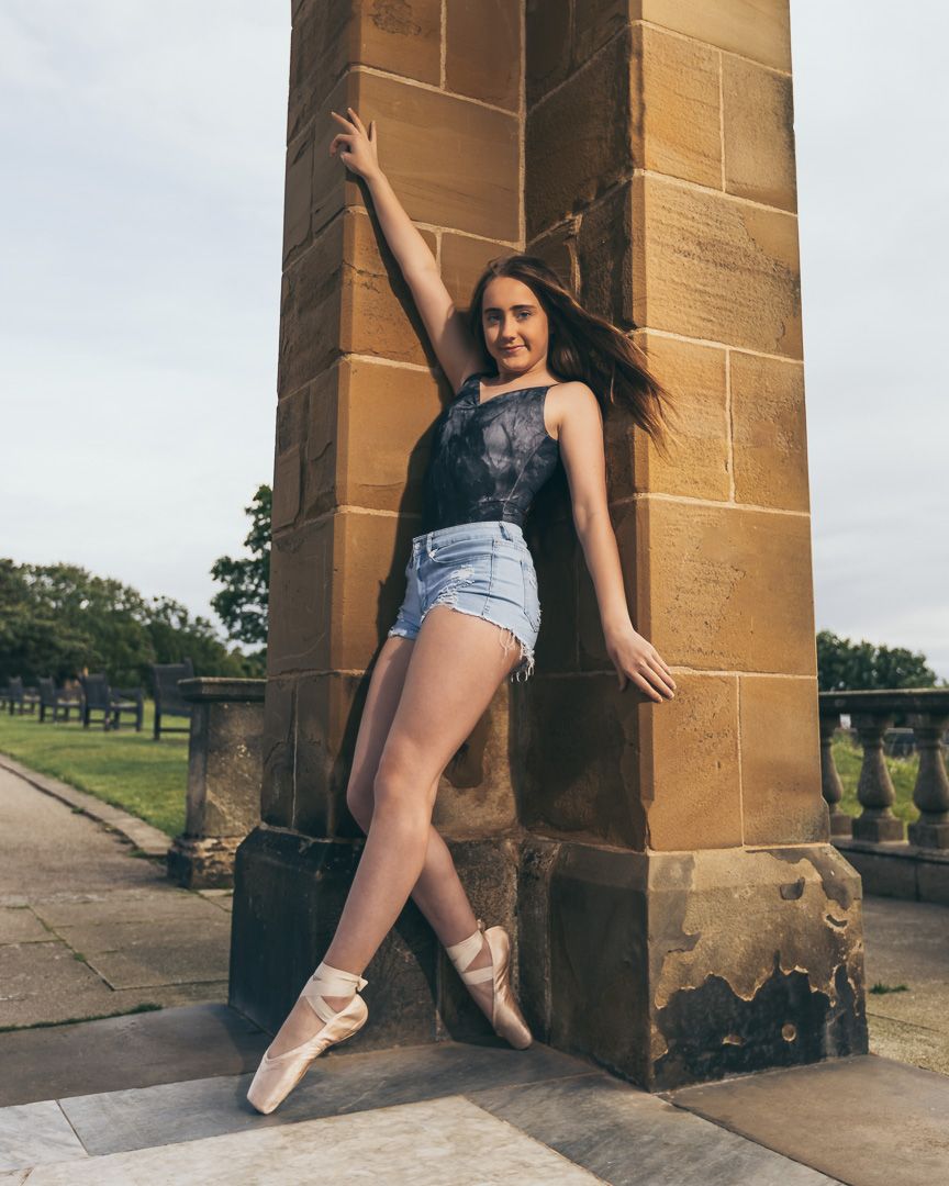 Ballet dancer in denim shorts poses gracefully against stone pillars in outdoor photoshoot series.