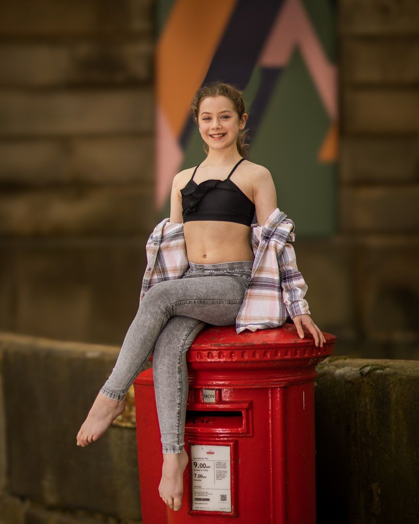 Young dancer posing playfully on red British post box in casual attire.