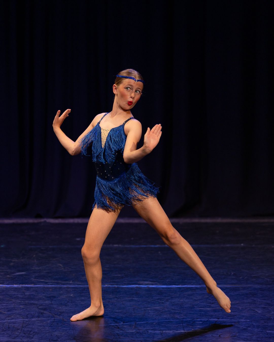 Dancer in a blue sequined costume performs a graceful split leap on a dark stage during a contemporary dance performance.