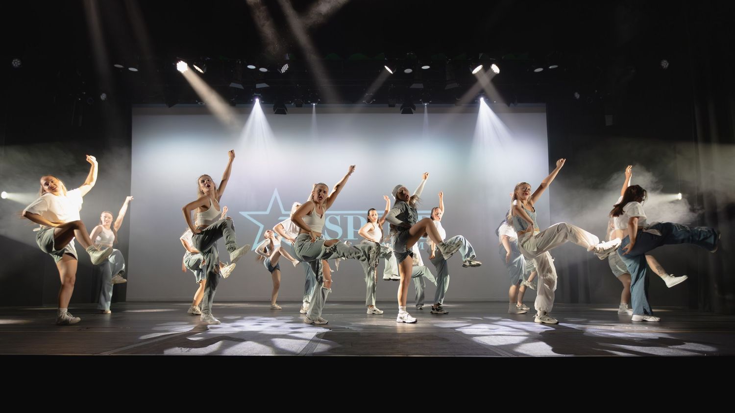 Energetic dance performers in coordinated costumes perform on a lit stage with dramatic spotlights and a star backdrop.