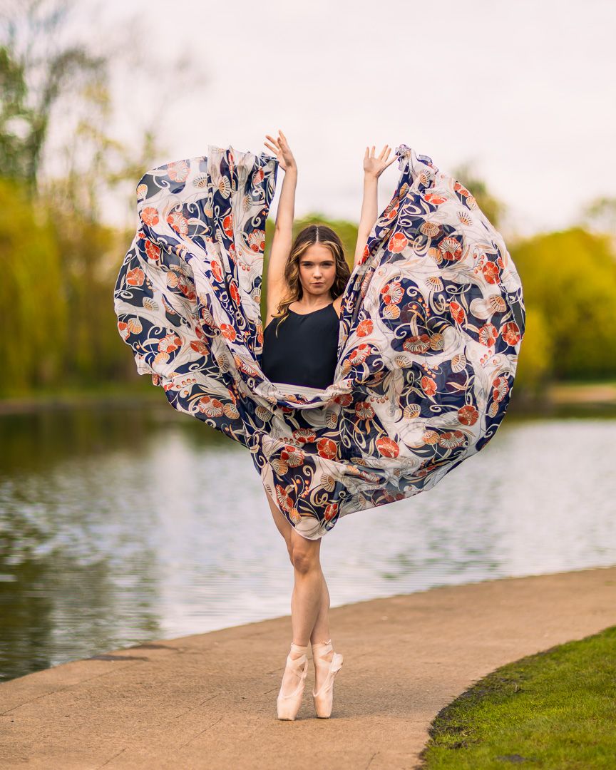 Dancer in floral wrap skirt performs ballet moves by lakeside at sunset.