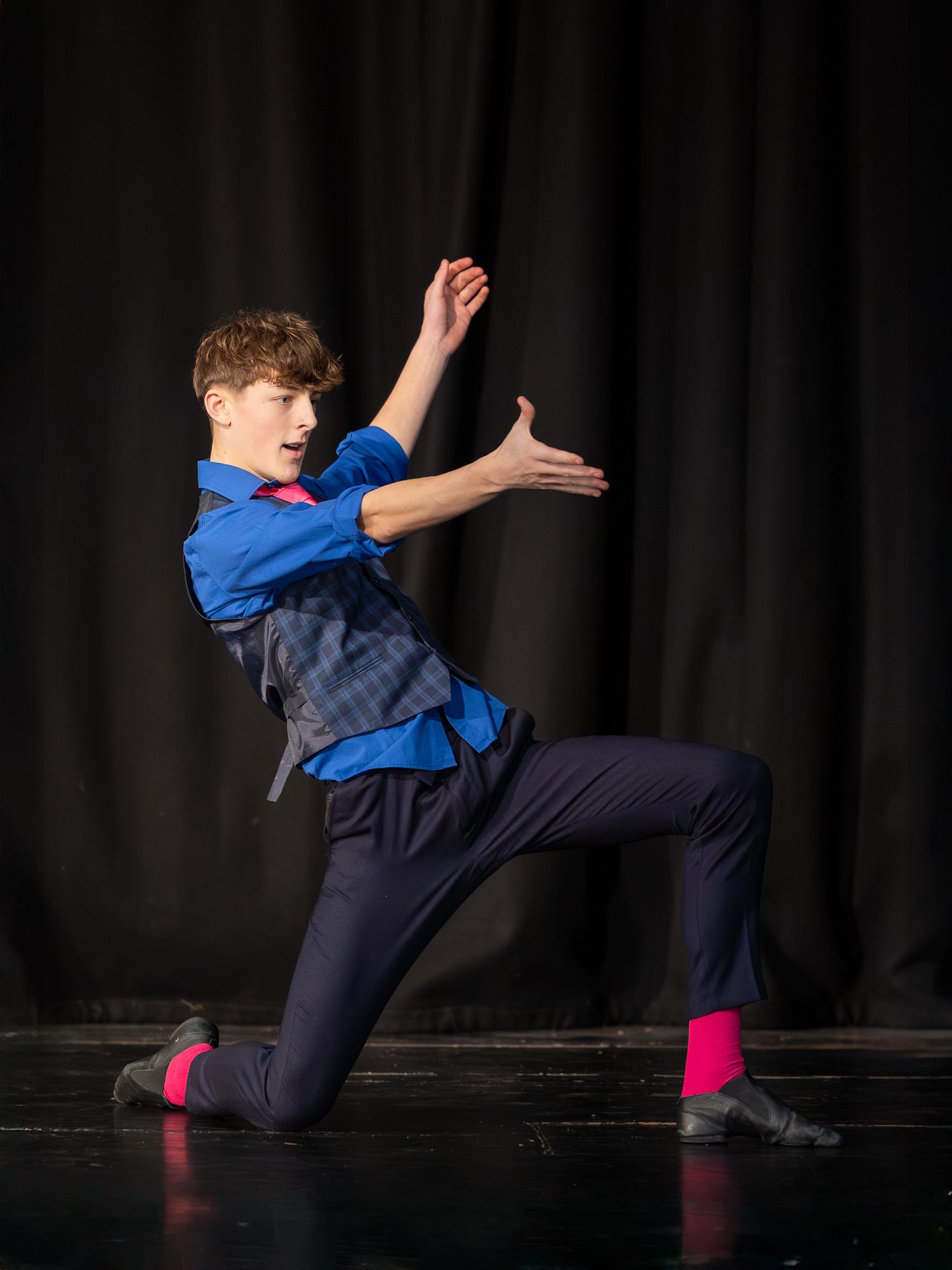 Young male dancer performing on stage in blue shirt, navy pants, and bright pink socks in a dramatic lunge pose.