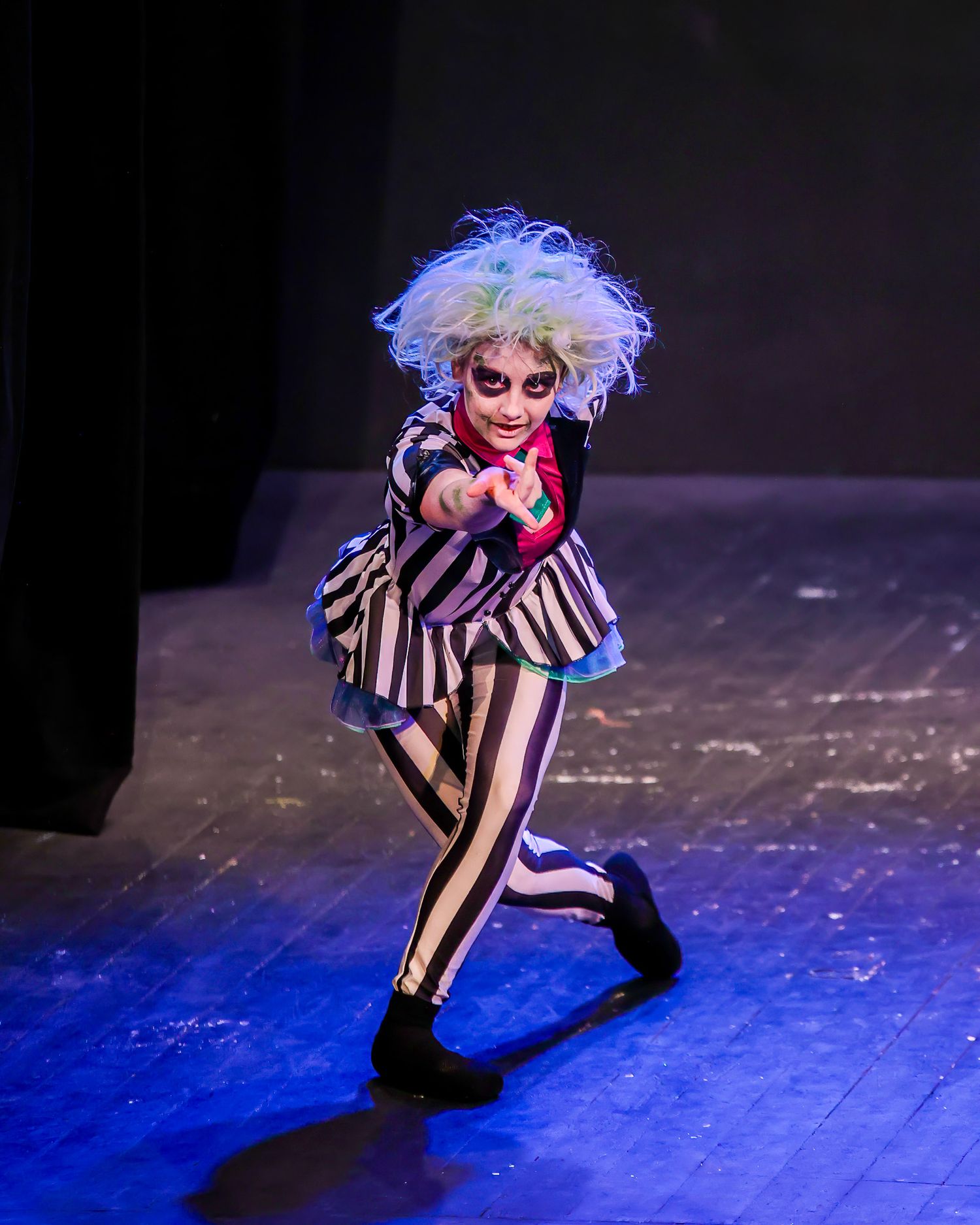 Child performer in Beetlejuice costume with wild gray wig dances on stage under colorful lights.