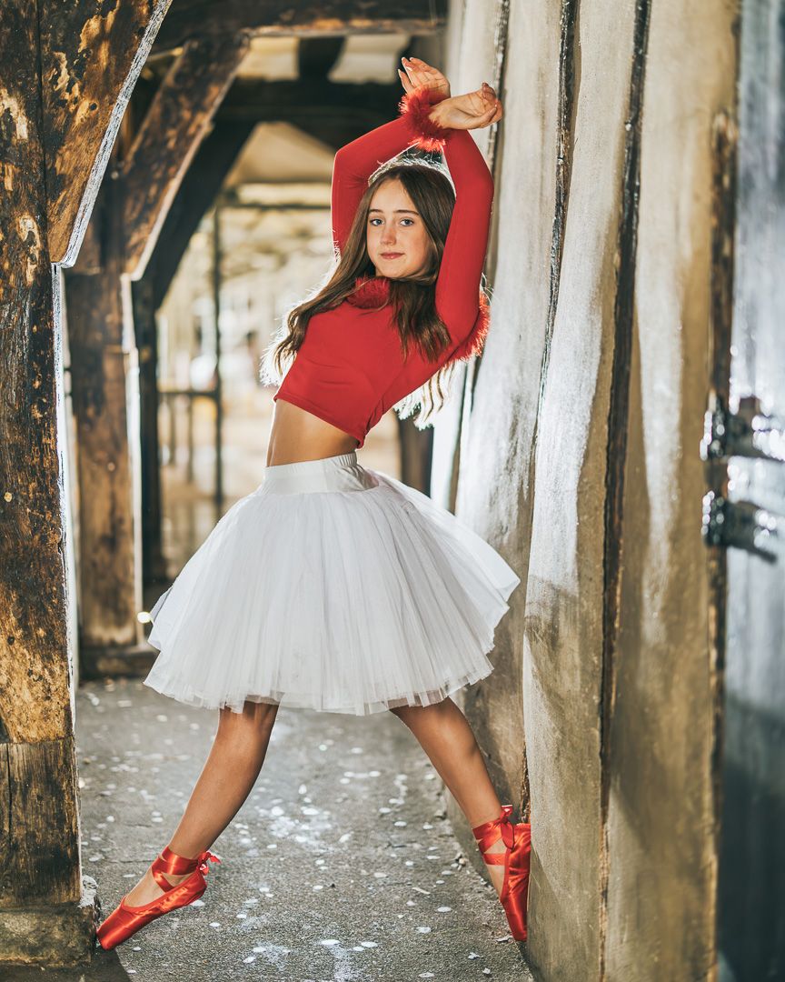 Young ballerina poses gracefully in white tutu skirt and red crop top with matching pointe shoes in rustic setting.