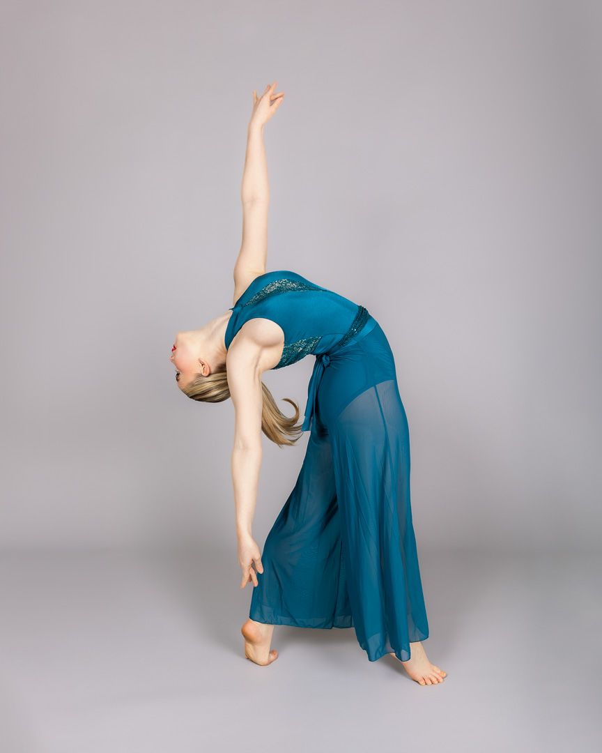 A dancer in a teal dress performs a dramatic backbend with one arm raised gracefully against a gray studio background.
