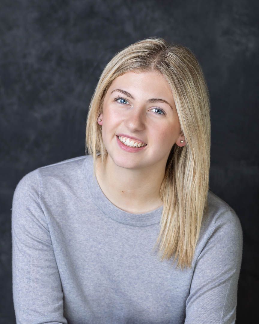 Smiling young blonde woman with blue eyes wearing a gray sweater poses for a professional portrait photo.