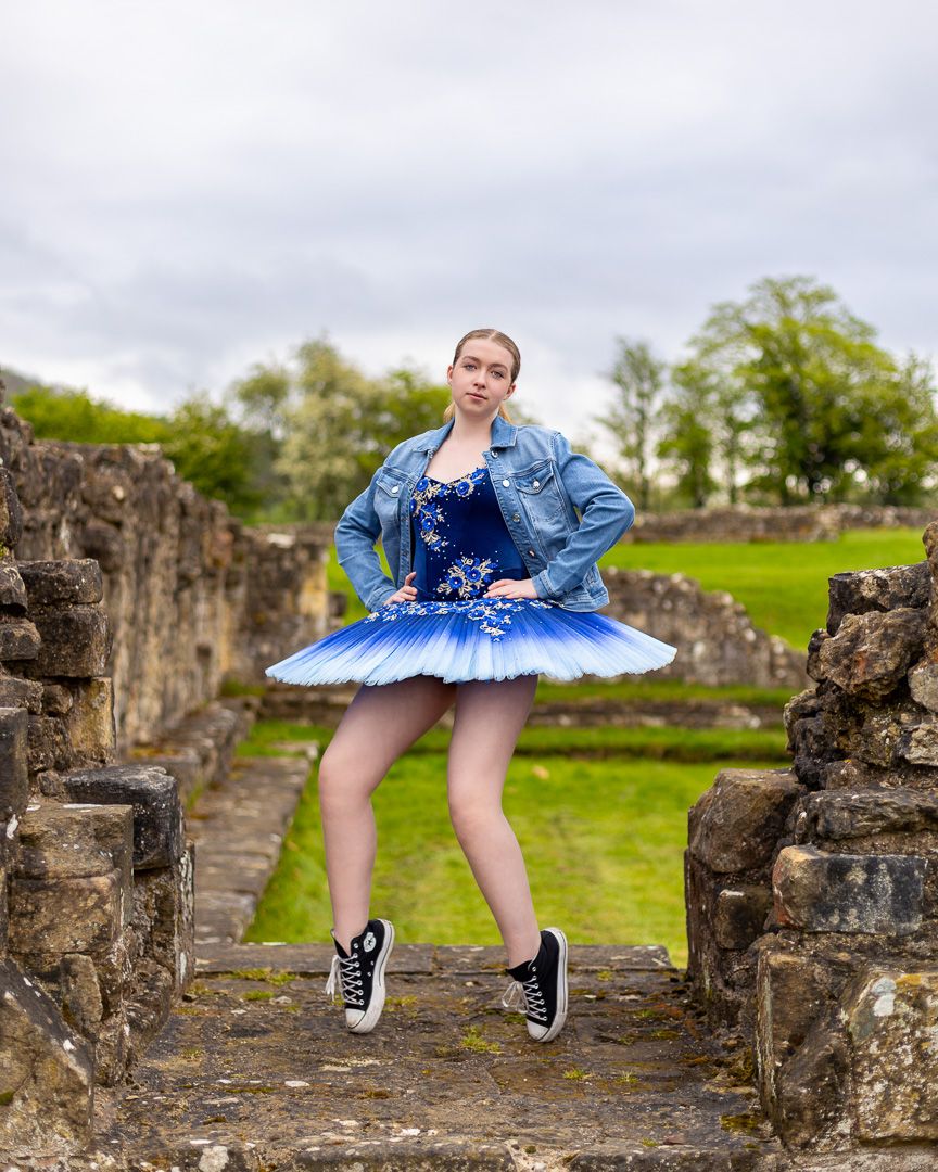 Young ballerina in blue tutu, denim jacket, and Converse sneakers poses gracefully among ancient stone ruins outdoors.