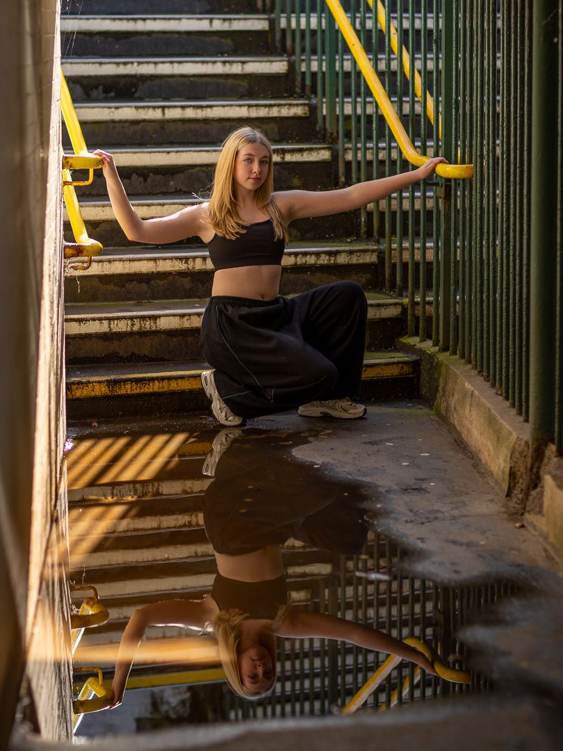 Young woman in black crop top and pants poses gracefully on stairs with yellow railings, reflected in puddle below.