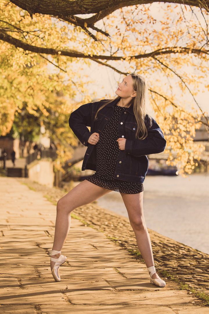 Ballet dancer in black jacket and polka dot dress performs on walkway under golden autumn trees.