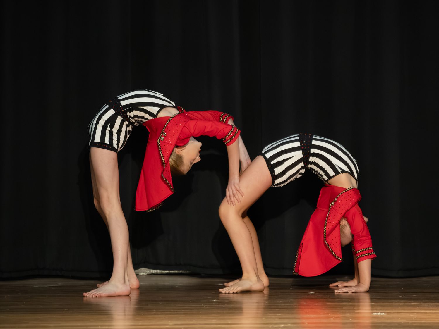 Two flexible acrobatic dancers in striped and red costumes perform a backbend routine on a wooden stage.