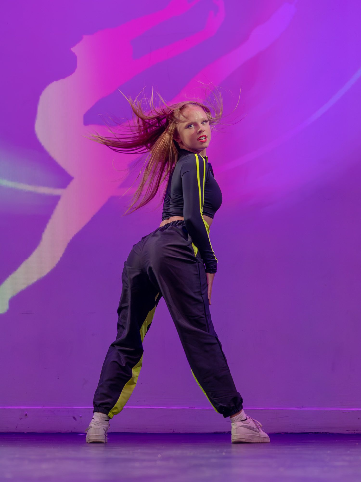 A young female dancer mid-move with hair flying, wearing black and yellow outfit against vibrant purple backdrop.