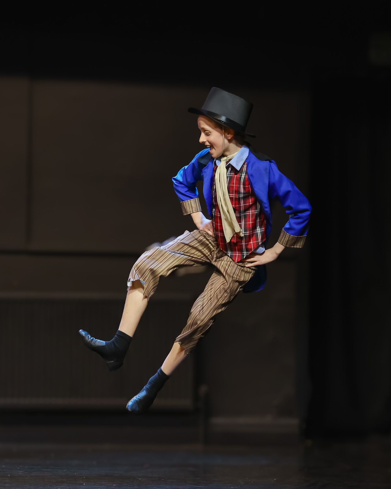 A young dancer leaps mid-air in a colorful costume with blue jacket, plaid shirt, striped pants, and top hat on stage.