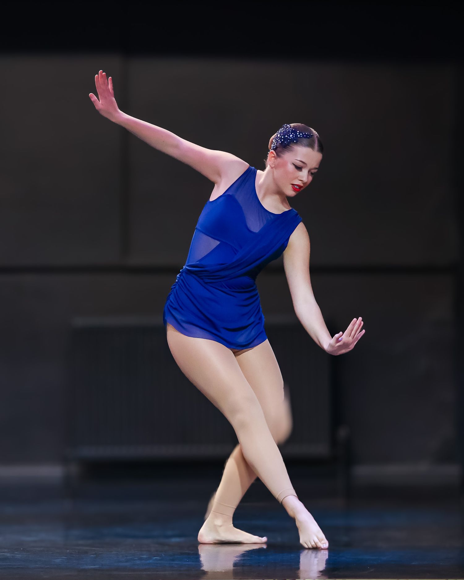 A dancer in a blue leotard performs an expressive contemporary dance routine on a dimly lit stage.