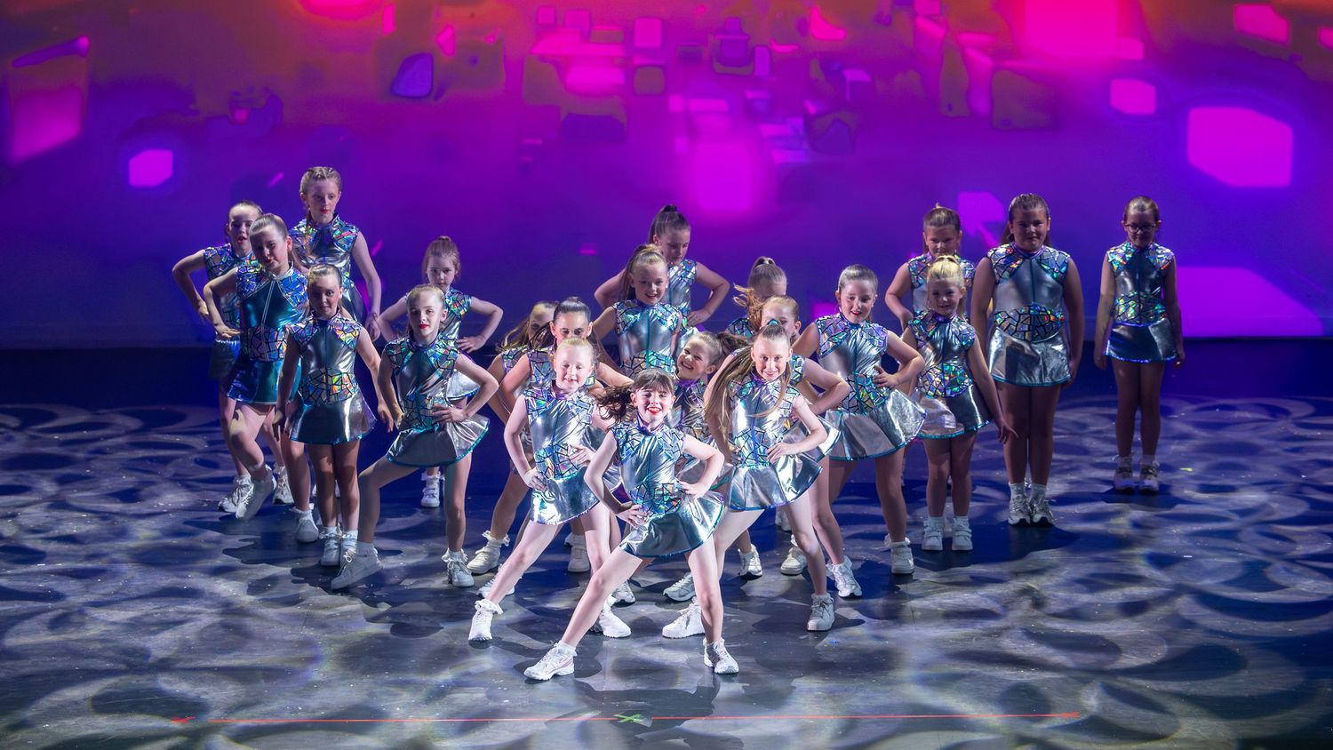 Young dancers in silver costumes perform on a colorful stage with purple and blue lighting.