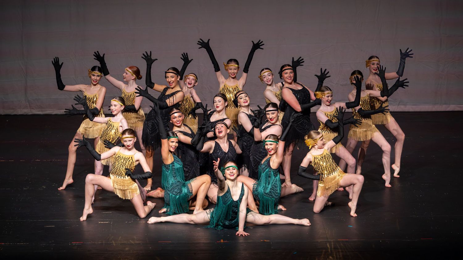 A large dance group poses on stage in gold and green flapper-style costumes with black gloves under dramatic lighting.