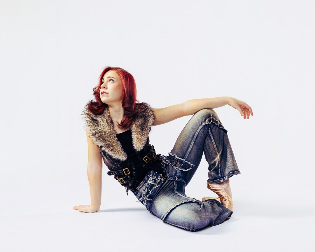 Red-haired woman in fur vest and jeans sitting on floor in a stylish pose against white background.