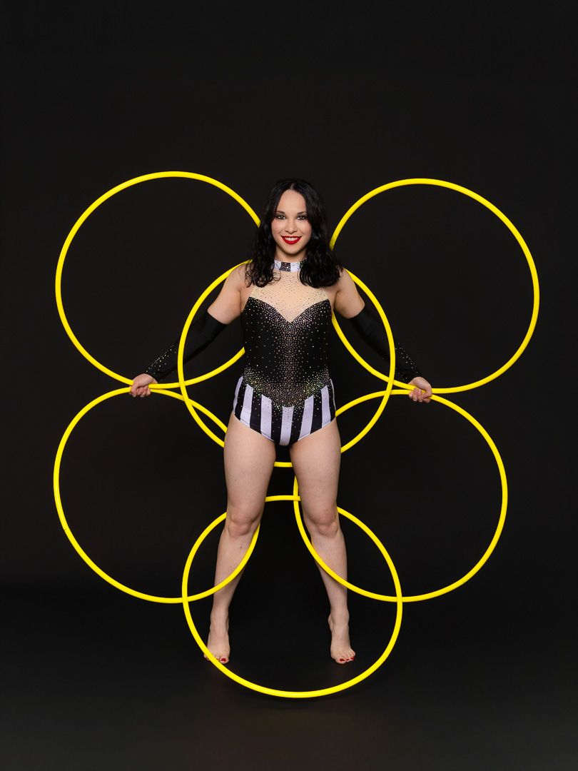 Performer in sparkly bodysuit poses with five yellow hula hoops arranged in a flower pattern against black background.