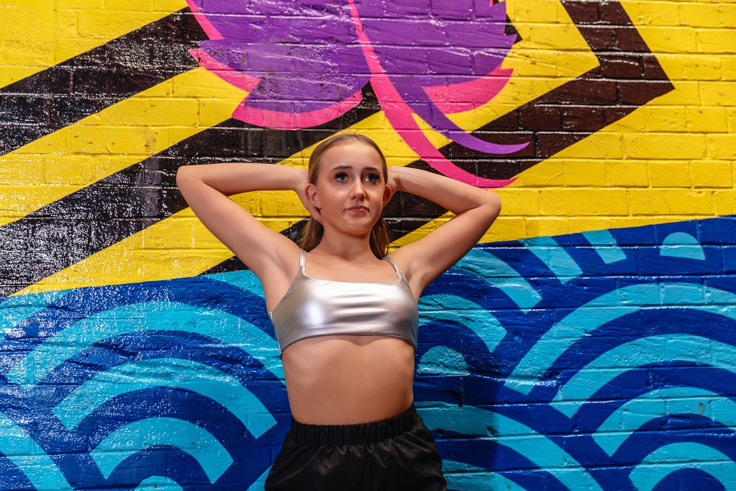 Young woman in silver crop top poses confidently against vibrant colorful graffiti mural wall.