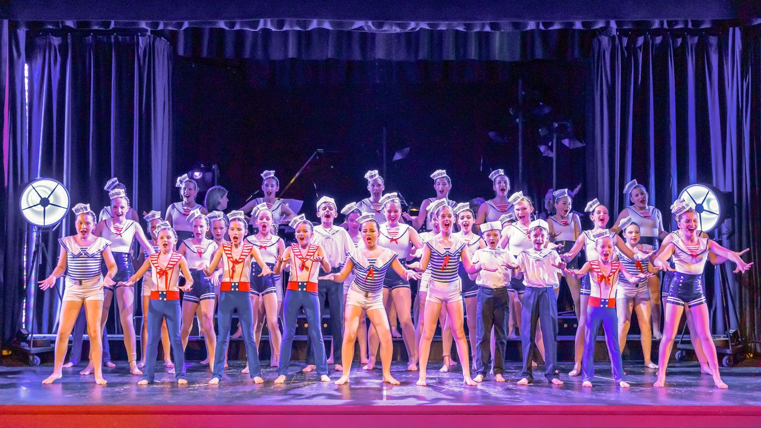 Large ensemble cast in colorful costumes performs on a theater stage with dramatic blue lighting.