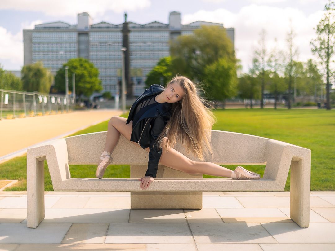 A flexible ballerina in a black jacket performs a split pose on a stone bench in a park.