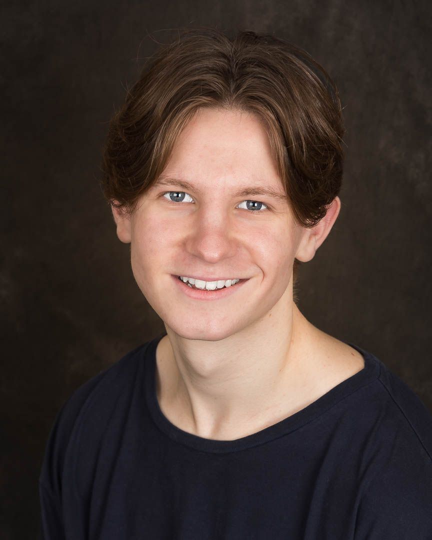 Young man with brown hair and blue eyes smiling warmly, wearing a navy shirt against a dark studio background.