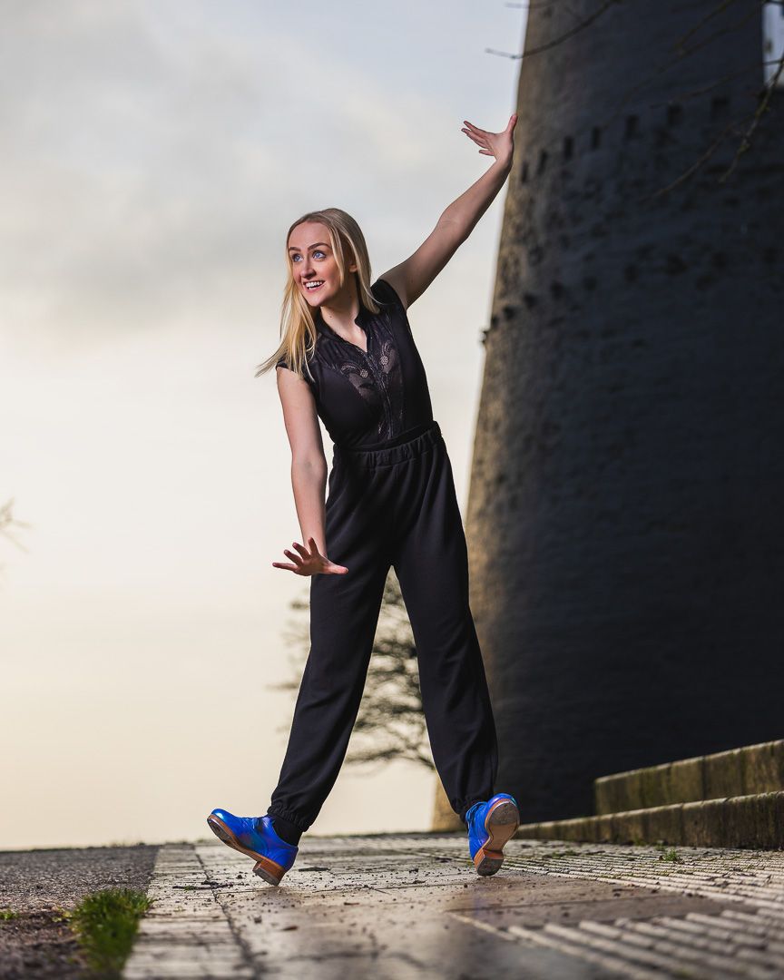 A person in a black jumpsuit and blue shoes poses joyfully against a lighthouse wall at sunset.