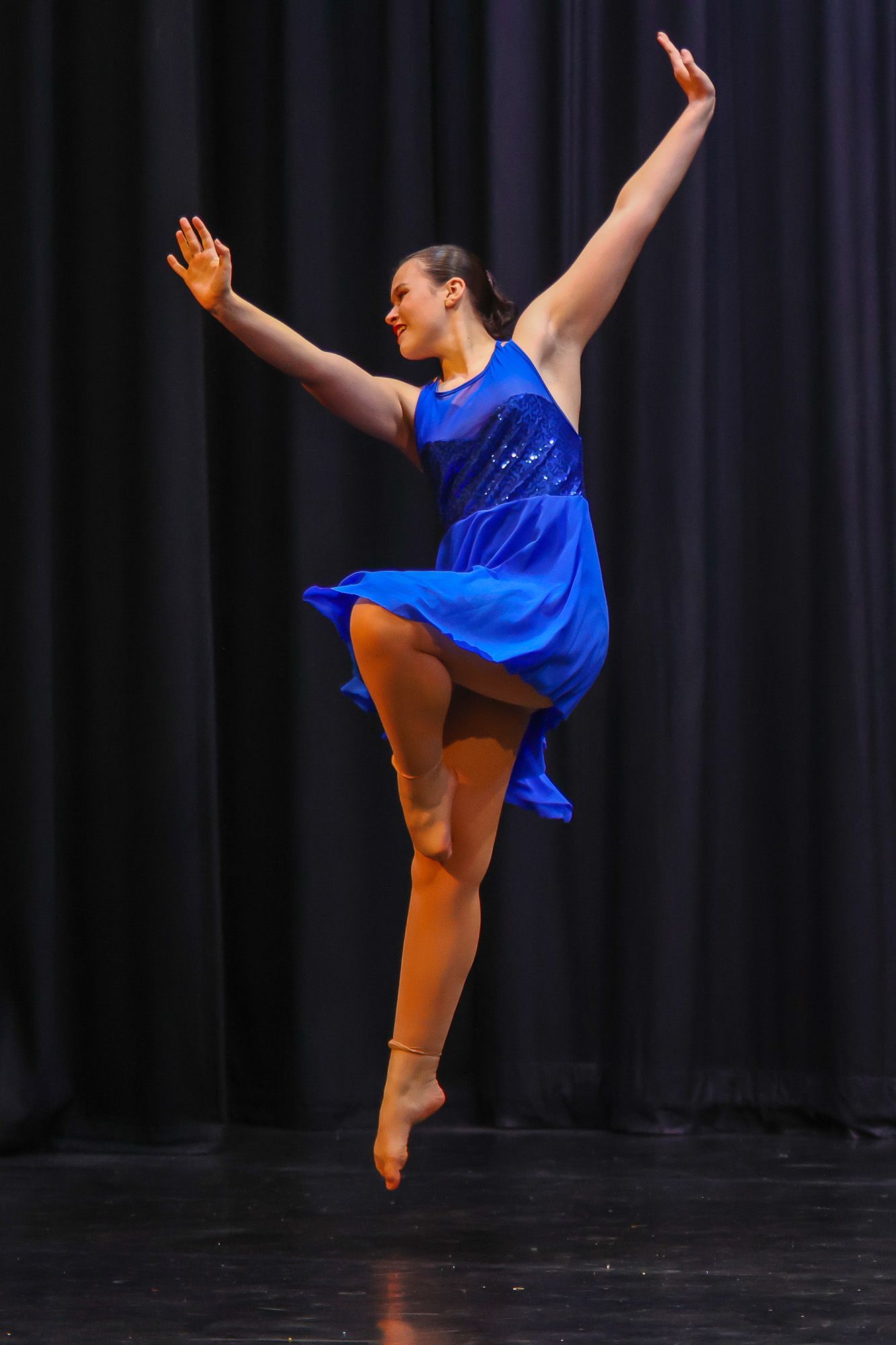 Female dancer leaps gracefully in blue dress on stage with arms extended.