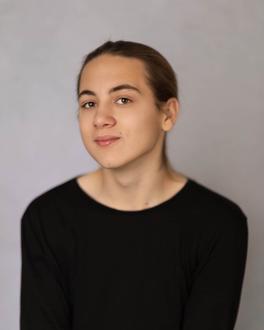 Young person with pulled-back hair wearing a black long-sleeve top, posing for a professional headshot photo.